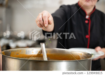 Close up of a chef in black uniform adding fresh herbs to a large pot of soup in a professional kitchen. Concept of gastronomy, seasoning, culinary preparation, and hospitality industry Close up of a chef in black uniform adding fresh herbs to a large pot of soup in a professional kitchen. Concept of gastronomy, seasoning, culinary preparation, and hospitality industry 129949815
