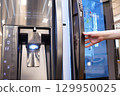 Close-up of a woman's hand holding a modern refrigerator, showcasing its sleek stainless steel surface and digital interface. 129950025