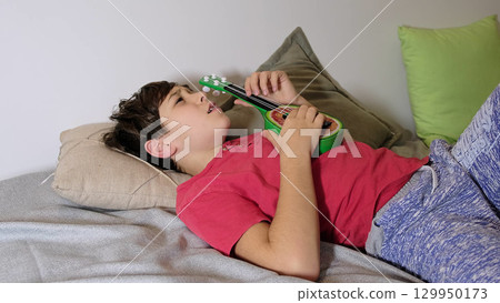 The boy gazes thoughtfully while lying down, embracing a moment of calm with his toy guitar. 129950173