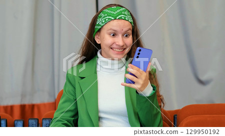 A candid capture of a woman in green sitting in an orange chair, absorbed in her phone. 129950192