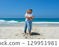 Couple standing barefoot on sandy beach with man hugging woman from behind in joyful moment. 129951981