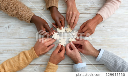 Hands of diverse people connecting puzzle pieces on wooden table, symbolizing teamwork and unity in warm, collaborative atmosphere 129953530