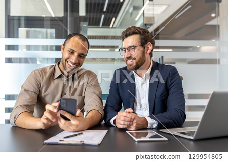 Two business professionals smiling while discussing work using a smartphone and tablet at their workplace. The modern office environment emphasizes teamwork, collaboration, and technology. Two business professionals smiling while discussing work using a smartphone and tablet at their workplace. The modern office environment emphasizes teamwork, collaboration, and technology. 129954805