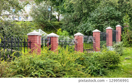 A wrought metal black fence with lattice and red brick posts separates the park from the lady on the hill behind the trees 129956991