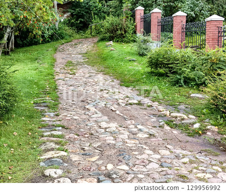 The rural cobblestone road runs along a wrought iron black metal fence with brick posts, in the middle of a park with grass and trees 129956992