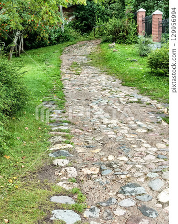 The rural cobblestone road runs along a wrought iron black metal fence with brick posts, in the middle of a park with grass and trees 129956994