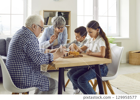 Grandparents teaching their little grandchildren to play chess at a table at home 129958577