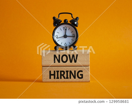 Now Hiring symbol. Wooden blocks with words Now Hiring. Beautiful orange background with alarm clock. Business and Now Hiring concept. Copy space. 129958681