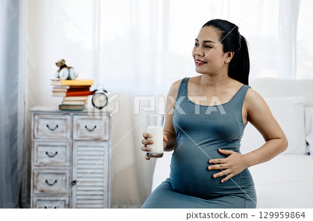 Pregnant woman in gray dress sitting on bed, smiling while holding glass of milk and touching belly at bedroom 129959864
