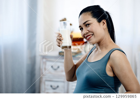 Smiling pregnant woman in gray dress holding glass of milk while sitting inside room with copy space 129959865