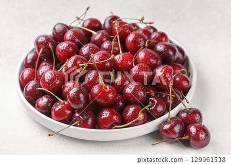 White plate full of fresh ripe red sweet cherry with water drops on white textured bakground White plate full of fresh ripe red sweet cherry with water drops on white textured bakground 129961538