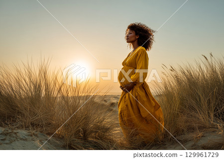 A pregnant woman is standing in a field of tall grass, wearing a yellow dress 129961729