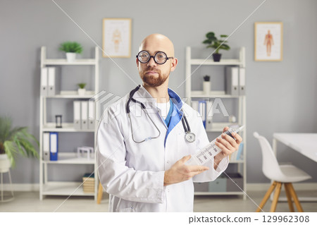 Funny doctor looking at camera holding big syringe in hands in medical clinic. Funny doctor looking at camera holding big syringe in hands in medical clinic. 129962308
