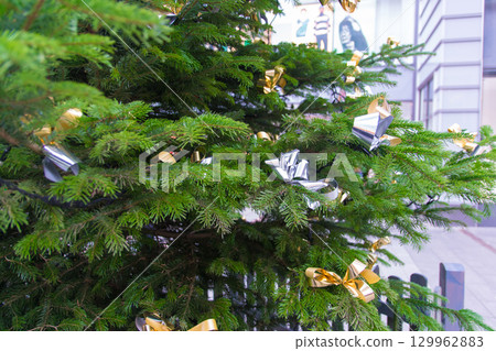 Festive street outdoor decor. Christmas tree with bright toys on city street background. Festive street outdoor decor. Christmas tree with bright toys on city street background. 129962883