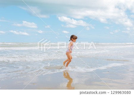 side view of little girl in the swimsuit running on the beach playing with waves 129963080
