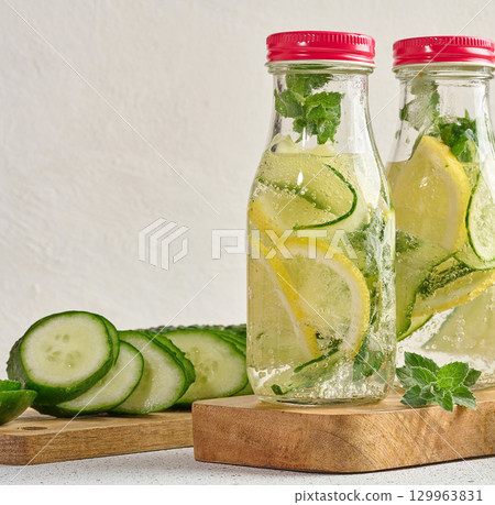 Glass bottles with red lids, filled with detox water containing cucumber and lemon slices and mint leaves, white table Glass bottles with red lids, filled with detox water containing cucumber and lemon slices and mint leaves, white table 129963831
