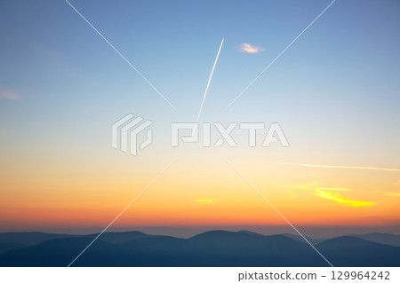Stunning sunset over mountain range with trails left by aircraft in clear blue sky 129964242