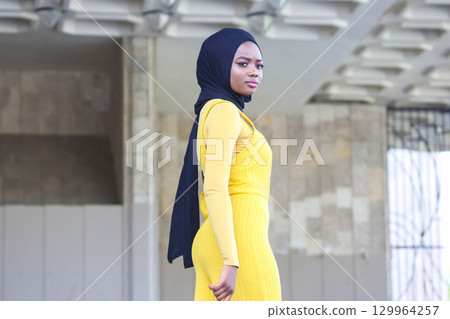 Portrait of an African woman in a yellow dress and black hijab in an urban setting 129964257