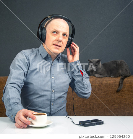Man enjoying coffee while listening to music with a cat nearby in a cozy indoor setting 129964260