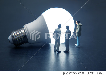 Miniature people gathered around a glowing light bulb in a dark environment Miniature people gathered around a glowing light bulb in a dark environment 129964365