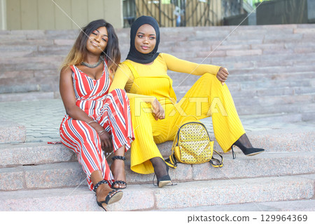 Two African women in stylish outfits relaxing on steps in a sunny urban setting 129964369