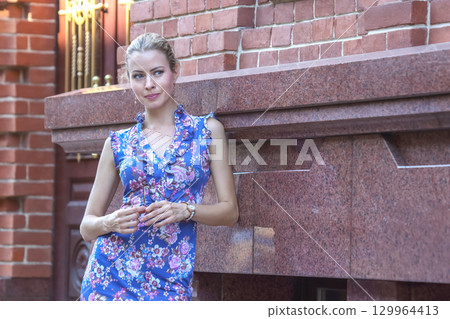 Woman in floral dress enjoying a moment outdoors near a brick wall in the afternoon sun 129964413