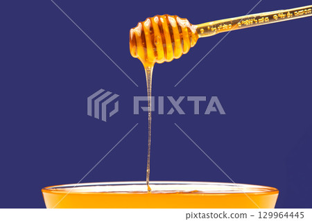 Golden honey drizzling from a wooden stick into a clear bowl against a vibrant background 129964445