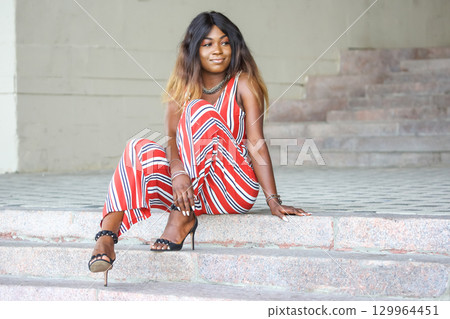 Striking African woman in stylish outfit sitting on steps during sunny day 129964451