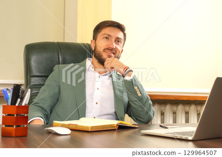 Professional man seated at desk with laptop and notebook in modern office setting 129964507