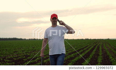 Agriculture, Rural Farmer Communicating, Man wearing a red hat, talking on mobile in a green field, casual day on the farm, relaxed stance, young farmer, fields stretching far, checking crop Agriculture, Rural Farmer Communicating, Man wearing a red hat, talking on mobile in a green field, casual day on the farm, relaxed stance, young farmer, fields stretching far, checking crop 129965072