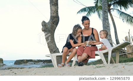 Mother and daughters sit on sunbed near sea petting their puppy and smiling on a sunny day Mother and daughters sit on sunbed near sea petting their puppy and smiling on a sunny day 129965870