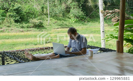 A young man relaxes in a hammock on a bamboo terrace, typing on his laptop, surrounded by lush greenery, tranquility of remote work in tropical setting  129965881
