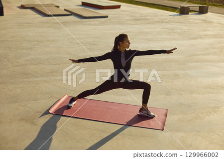 Fit young woman doing Warrior pose on yoga mat during outdoor summer workout in city Fit young woman doing Warrior pose on yoga mat during outdoor summer workout in city 129966002