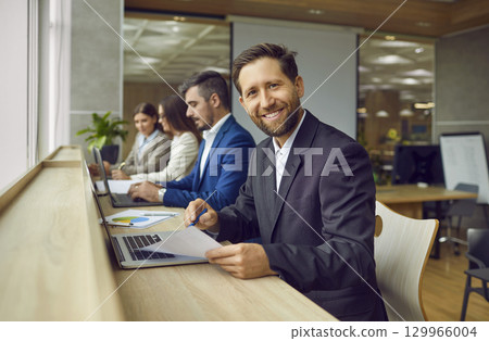 Male employee sitting in modern office at same desk with coworkers and looking at camera. 129966004