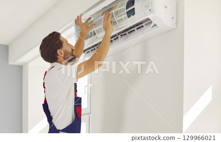 Male professional technician cleans or replaces air conditioner filter hanging on wall inside house. Male professional technician cleans or replaces air conditioner filter hanging on wall inside house. 129966021