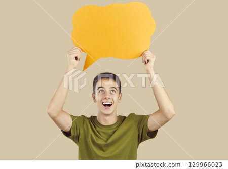 Happy young man holding orange speech balloon or thinking bubble on beige background 129966023