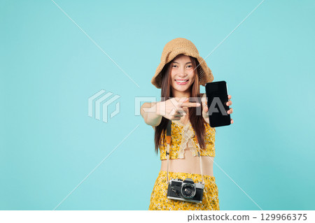 Smiling asian young woman traveler showing phone blank screen points, wearing summer floral outfit and holding mobile phone up while presenting its features, headshots isolated studio blue background 129966375
