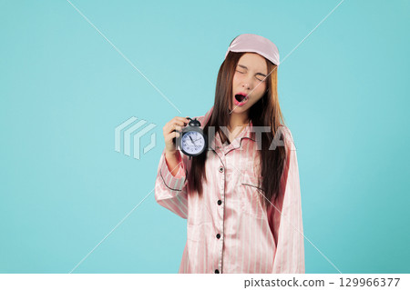 Tired young Asian woman awake time holding clock in pajamas, just woken from sleep with a sleep mask, shocked by the alarm, she needs more sleep now, headshots isolated studio blue background 129966377