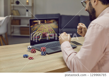 Young man sitting at the desk and playing gambling in online casino at home. 129966785