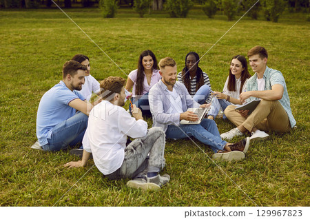 Diverse team of happy business students working together, sitting on grass in park and using laptop 129967823