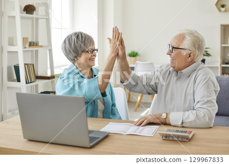 Elderly couple sitting at the table with laptop and bills giving high five each other at home. 129967833