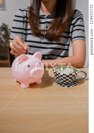 A woman places a coin in a piggy bank while enjoying a matcha latte, symbolizing savings and relaxation. 129972772