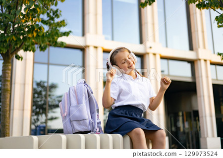 A Caucasian student girl in a schoolyard, wearing headphones, enjoying music, with a school building in the background. 129973524