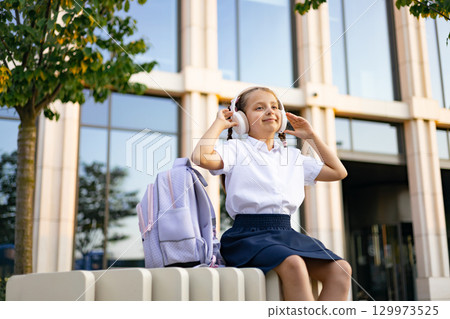 Caucasian schoolgirl in a schoolyard wearing headphones and listening to music, with a backpack. Caucasian schoolgirl in a schoolyard wearing headphones and listening to music, with a backpack. 129973525