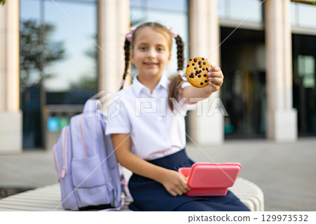 A Caucasian schoolgirl offers a chocolate chip cookie in the schoolyard, with a backpack and lunchbox. A Caucasian schoolgirl offers a chocolate chip cookie in the schoolyard, with a backpack and lunchbox. 129973532