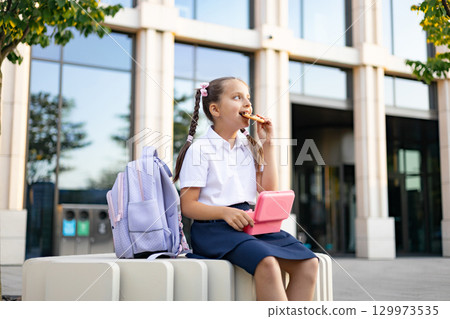 Caucasian schoolgirl with pigtails eats a snack while sitting near a school building with a backpack. 129973535