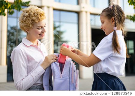 A mother helps her daughter pack her lunchbox in the schoolyard, preparing for the school day. 129973549