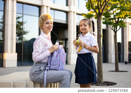 A mom and her school-aged daughter are outside of the school building, smiling and holding snacks. A mom and her school-aged daughter are outside of the school building, smiling and holding snacks. 129973560