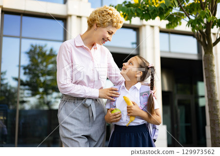 A mom and her student daughter smile at each other in front of a school building, ready for the day. A mom and her student daughter smile at each other in front of a school building, ready for the day. 129973562