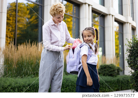 A mother helps her daughter pack her backpack in front of a school building. The little girl is wearing her school uniform. 129973577